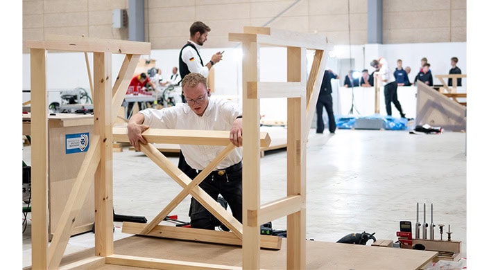 Zwei Männer arbeiten in einer Werkstatt an einem Holzregal; einer prüft das Regal, der andere nutzt ein Smartphone im Hintergrund.