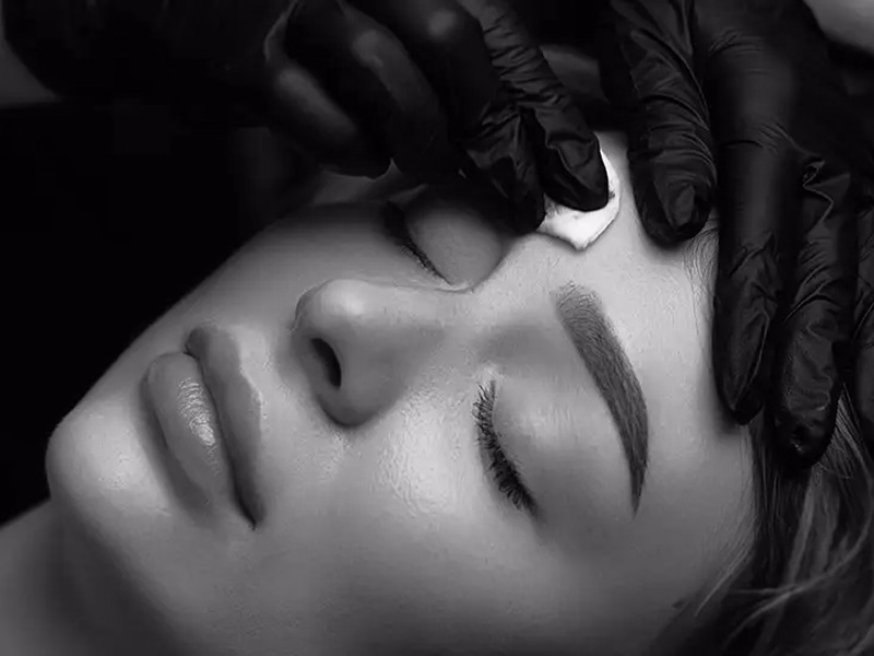 Detaillierte Gesichtspflege nach Microblading und Microshading, bei der jemand mit schwarzem Handschuh ein Pad auf die Stirn legt.
