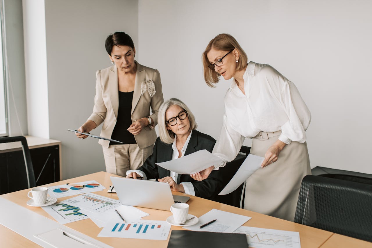 a-woman-in-white-dress-shirt-showing-paper-to-woman-sitting-at-a-table-with-laptop-7433855 A Woman in White Dress Shirt Showing Paper to Woman Sitting at a Table with Laptop