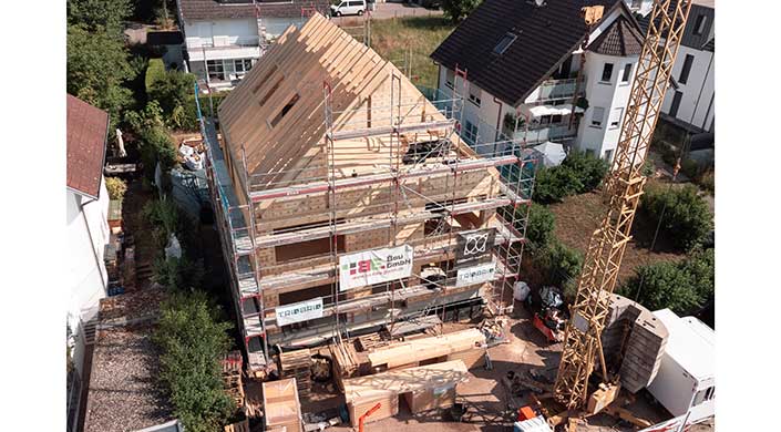 Ein Wohnhaus im Rohbau mit Holzarbeiten und Baugerüst, aufgenommen bei Tageslicht. Baumaterialien und Werkzeuge sind rund um die Baustelle verteilt.