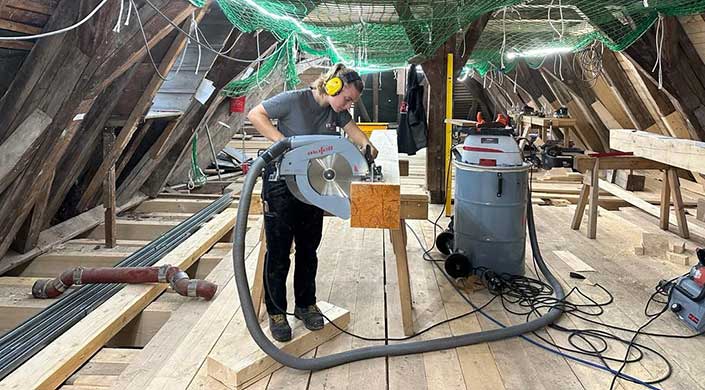 Person mit Gehörschutz benutzt eine Kreissäge, um ein großes Holzbrett auf einer Werkbank in einer Werkstatt zu schneiden.
