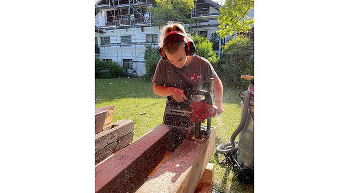 Eine Person trägt Gehörschutz und arbeitet mit einer elektrischen Säge an einem Holzbalken im Freien, während Sägespäne fliegen.