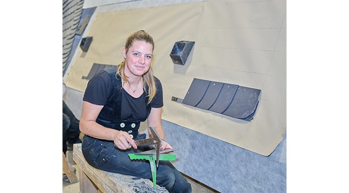 Eine Frau sitzt mit Werkzeugen vor einer Wand mit geometrischen Formen aus Metall und Stein, während sie in die Kamera lächelt.