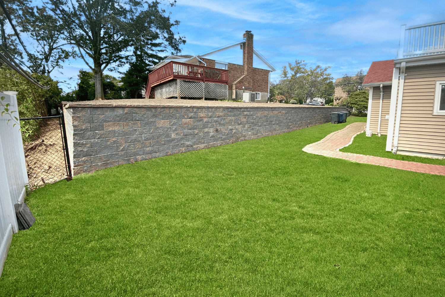 A backyard with a green lawn, Retaining Walls made of stone, a chain-link fence, and a brick pathway alongside the house. A raised deck is visible behind the wall.
