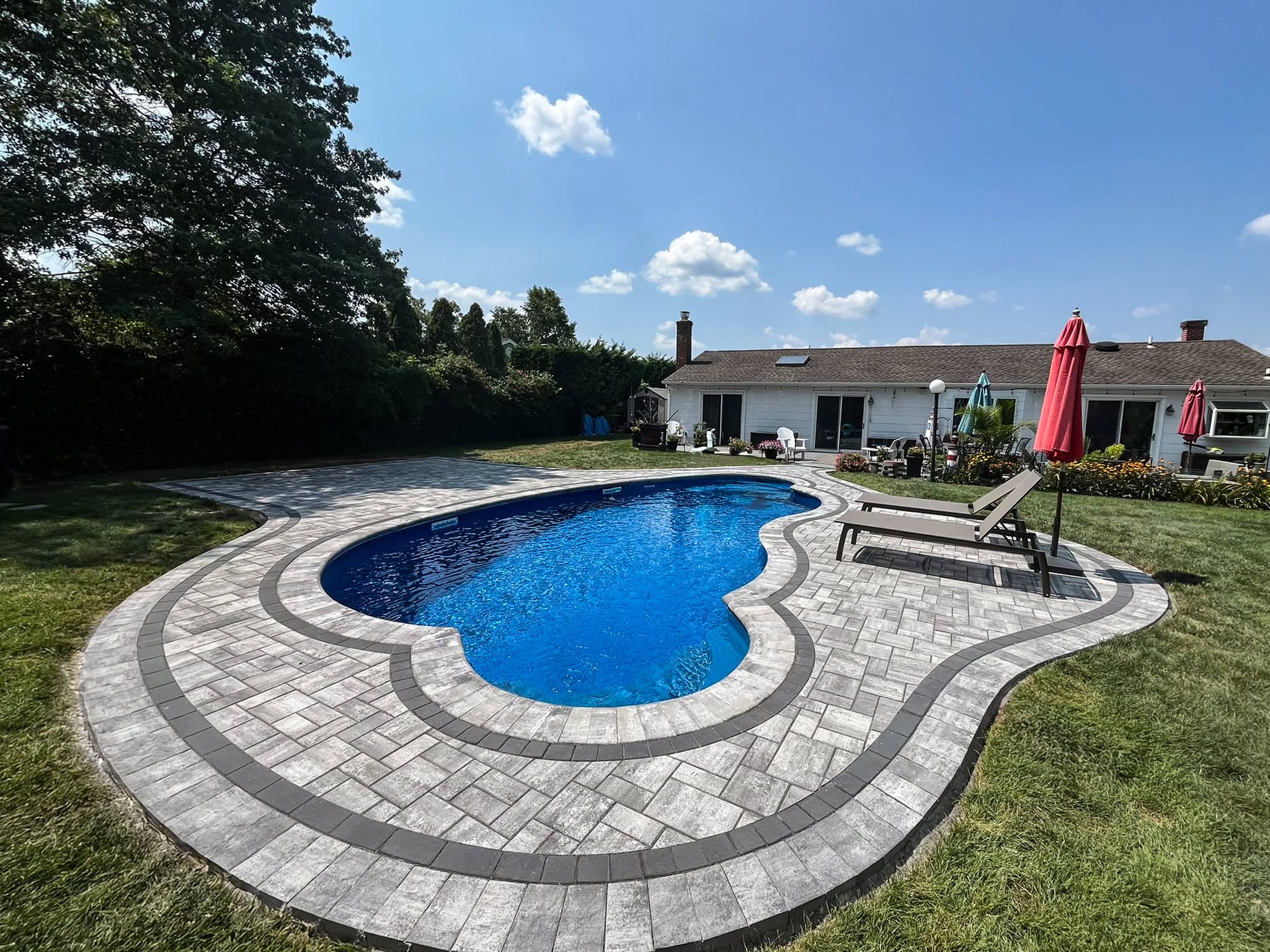 Backyard with an in-ground, kidney-shaped pool surrounded by stone pavers, lounge chairs, and red umbrellas next to a single-story house under a partly cloudy sky.