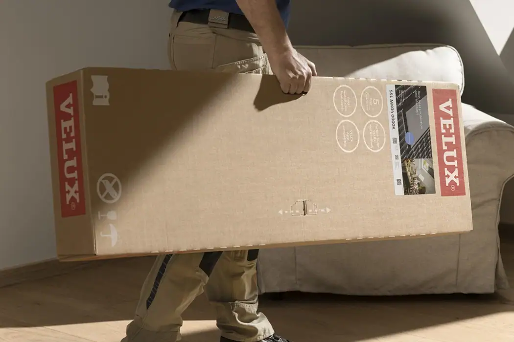 Eine Person trägt einen großen, rechteckigen Karton mit dem VELUX-Logo vor einem Sofa in einem hellen Raum.