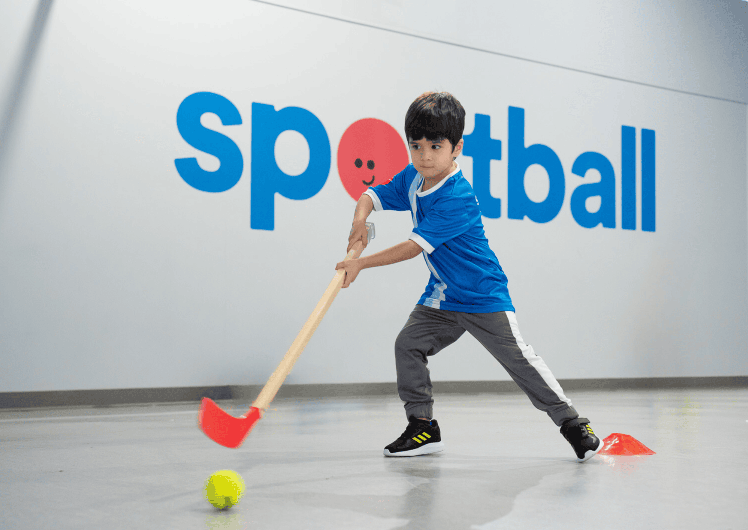 Young child in a Sportball uniform plays floor hockey during an indoor class with a training stick and ball.