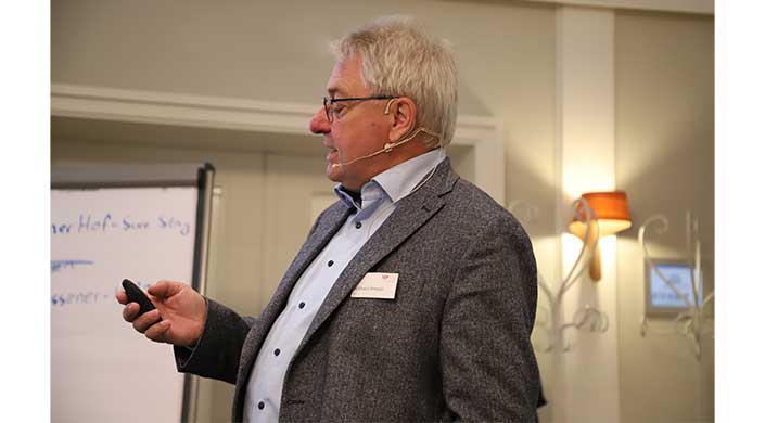 Bild von Eckhard Rimpel vom Institut für Ziegelforschung Ein älterer Mann mit Brille und grauem Haar trägt ein Mikrofon-Headset und einen Anzug, steht seitlich und spricht bei einer Veranstaltung.