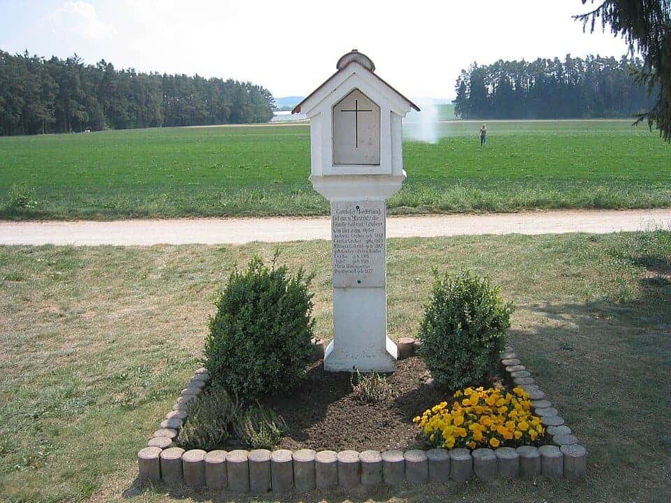 A white wayside shrine with a small roof and a cross, with an inscribed plaque, between two shrubs and yellow flowers, beside a gravel track and green fields