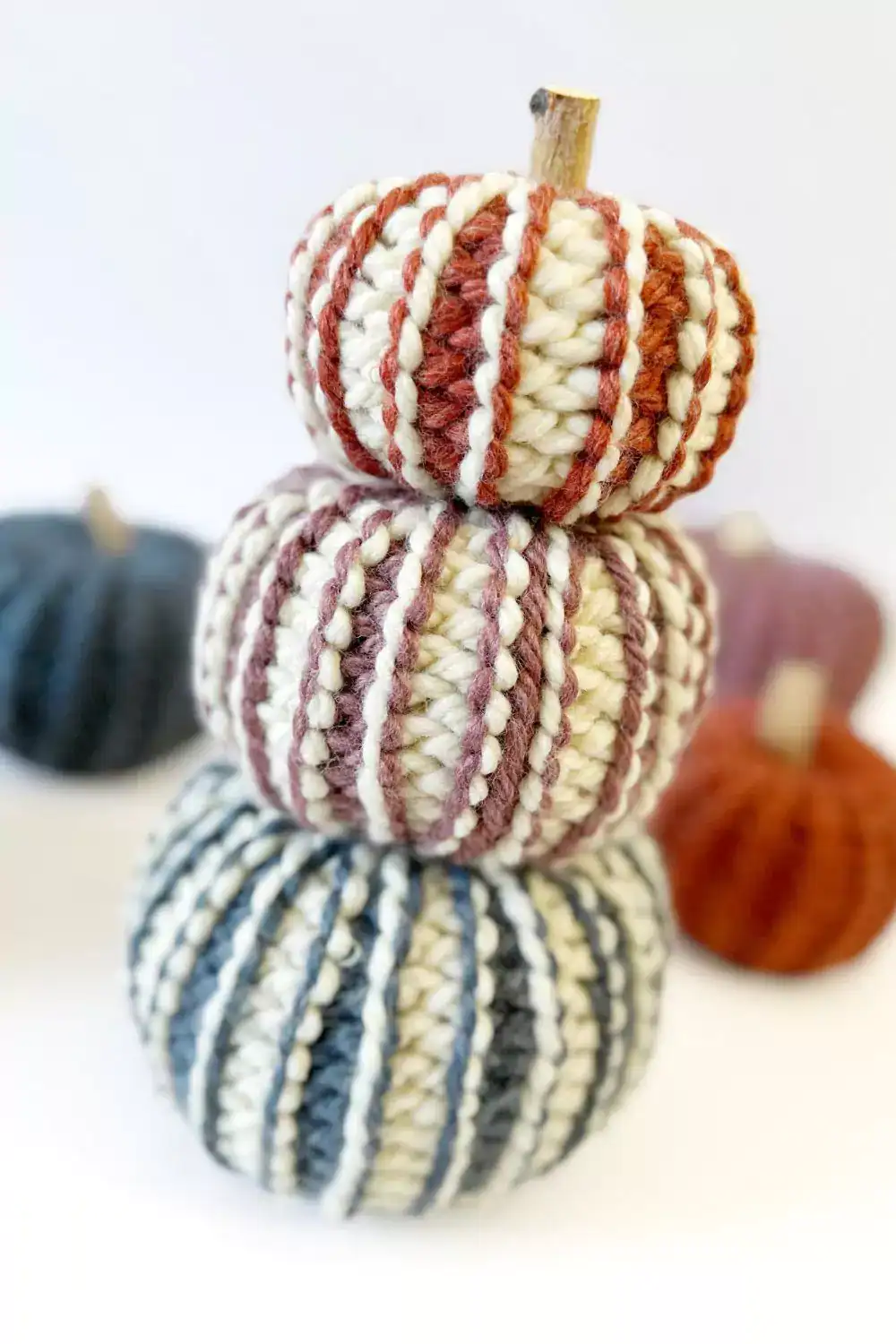 Three striped, chunky ribbed crochet pumpkins stacked on a white surface; two more blurred in the background, no visible text. Free crochet pattern