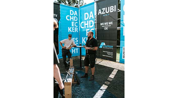 Ein Mann steht im Freien vor blauen Werbebannern, trägt schwarze Arbeitskleidung und spricht mit einer anderen Person an einem Stehtisch.