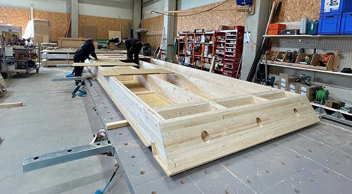 Großes Holzbauteil liegt in einer Werkstatt, mit Regalen und Werkzeugen im Hintergrund.