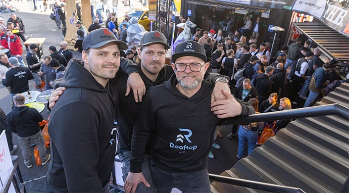 Zwei Männer mit Brillen und Mützen stehen nebeneinander, tragen schwarze Hoodie mit „Rooftop“-Logo, Menschenmenge und Ausstellungsstücke im Hintergrund.