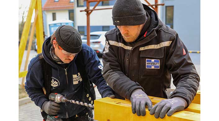 Zwei Personen arbeiten draußen zusammen an einem gelben Holzobjekt; sie tragen dicke Jacken und Mützen, im Hintergrund sind Autos und Gebäude sichtbar.