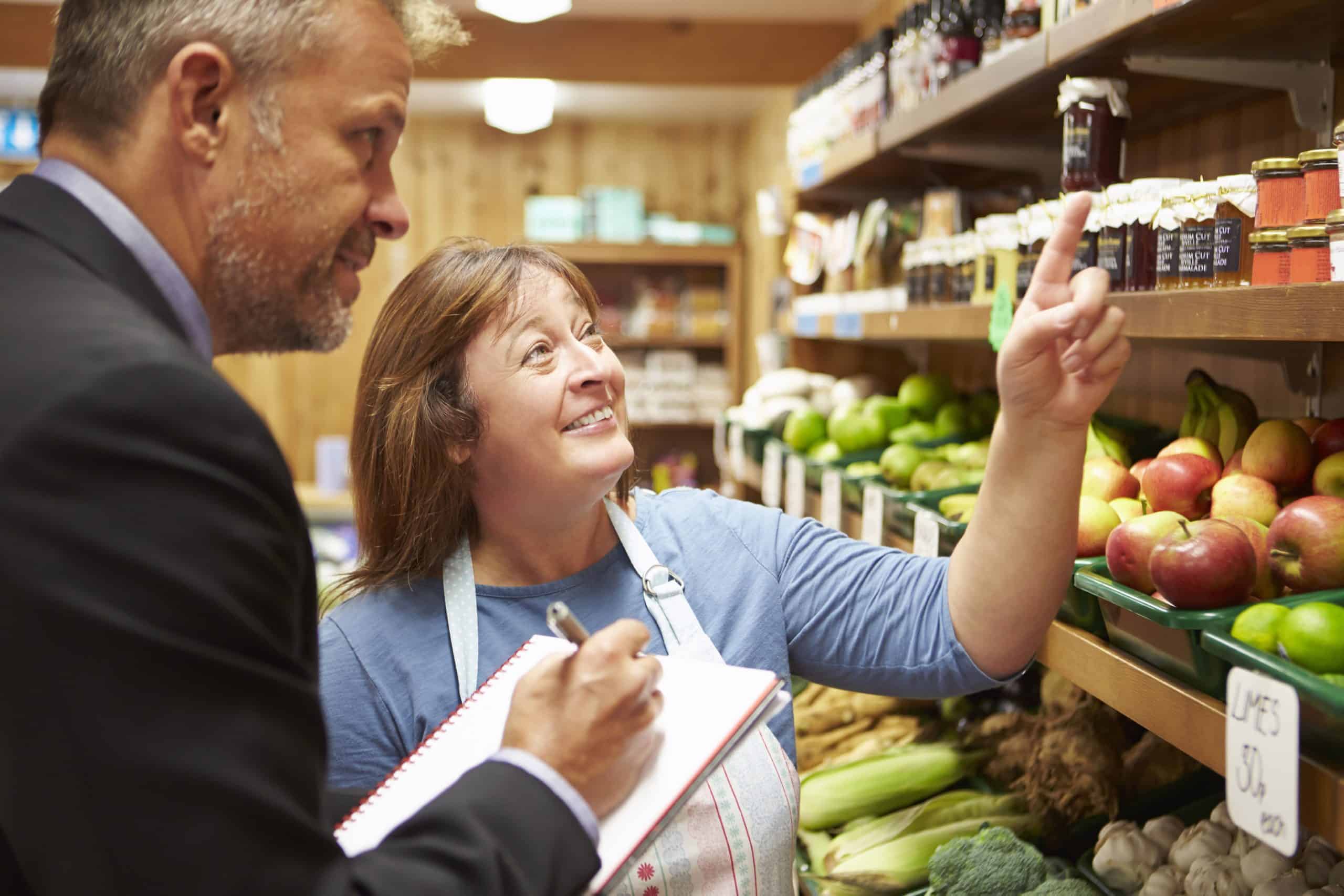 A woman in an apron points at jars on a grocery shelf, while a man in a suit takes notes on a clipboard. Fresh produce is displayed beside them.