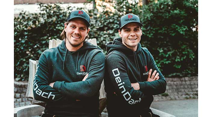 Zwei junge Männer stehen Rücken an Rücken, tragen dunkle Hoodies und Baseballcaps, im Freien vor grünem Hintergrund, beide lächeln leicht.
