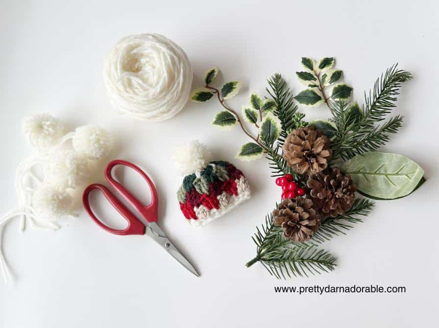 Craft supplies on a white surface: white yarn, pom-poms, red scissors, a small crocheted hat, pinecones, berries, and greenery. Free crochet mini hat christmas ornament pattern