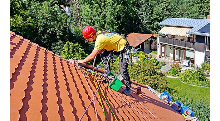 Ein Arbeiter mit Helm und Sicherheitsausrüstung arbeitet auf einem roten Ziegeldach, umgeben von Seilen, Bäumen und einem Haus im Hintergrund.