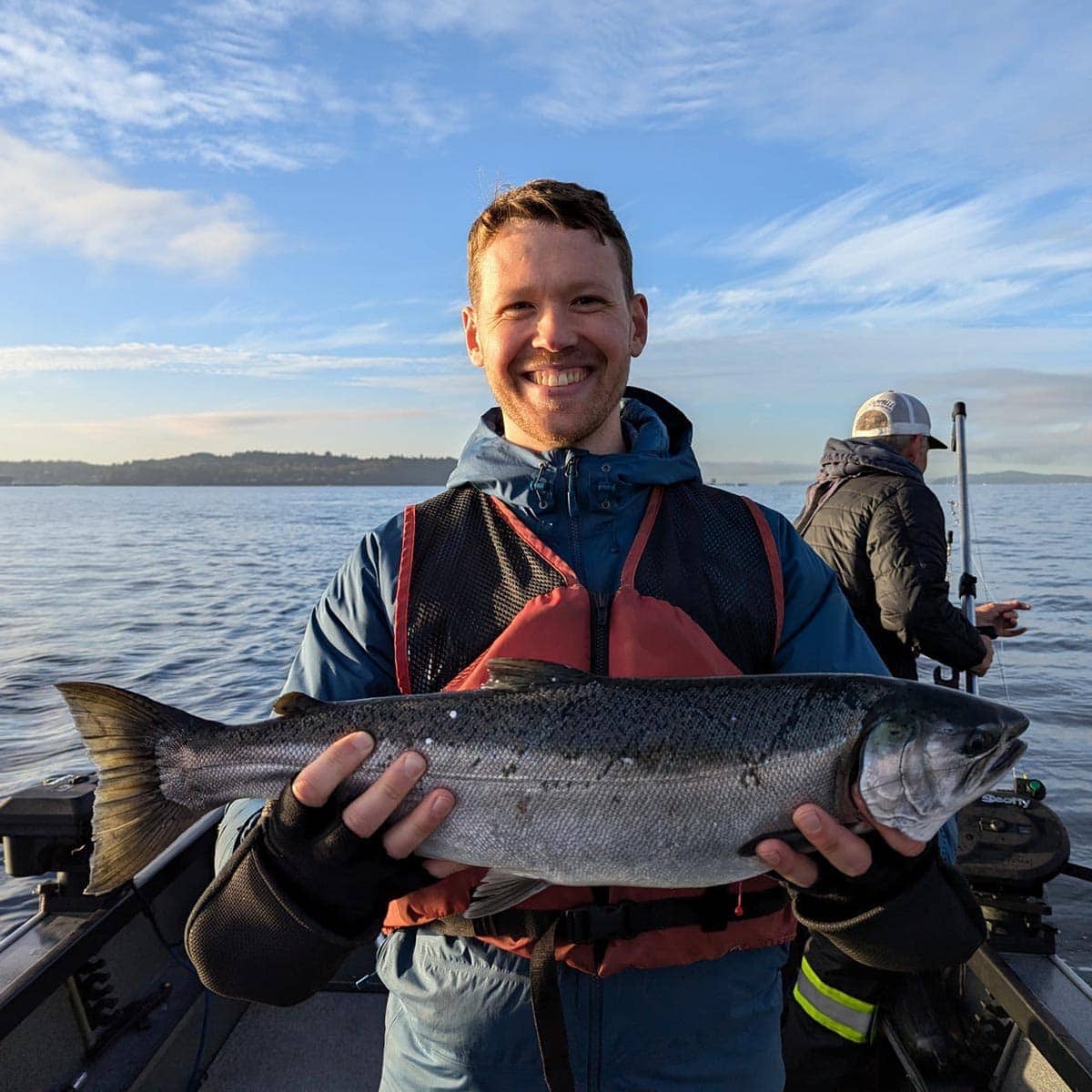 A man standing on a boat holds a large fish with both hands, smiling at the camera. Another person is fishing in the background on a calm body of water.