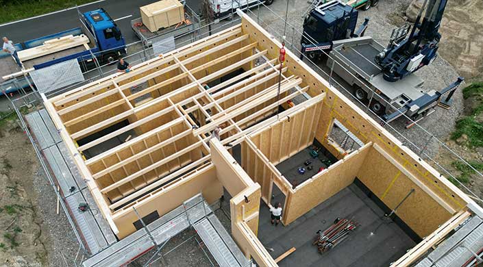 Holzrahmenbau eines Gebäudes, sichtbare Dachbalken und Wände, Bauarbeiter arbeiten an der Konstruktion auf einer Baustelle, Gerüst am Rand.