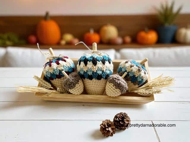A wooden tray holds cream, blue, and brown crocheted acorns with wheat stalks on a white table; blurred pumpkins in background. Free crochet pattern