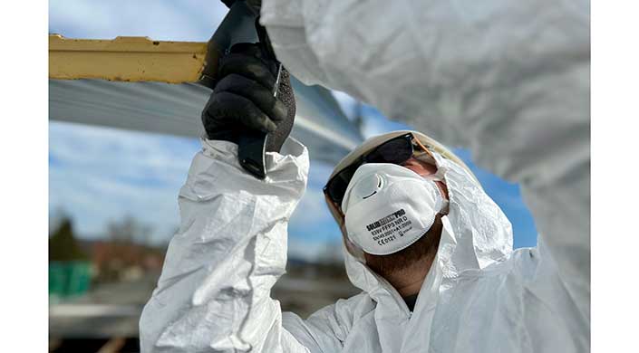 Person mit Schutzanzug, Maske und Schutzbrille arbeitet im Freien, hält ein schwarzes Werkzeug, blauer Himmel im Hintergrund.