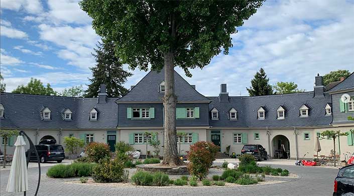 Ein großes Haus mit dunkelgrauem Dach und grünen Fensterläden, davor steht ein hoher Baum im Vorgarten.