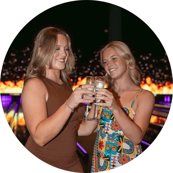 Two women enjoying drinks at a luxury bowling alley with vibrant lighting.
