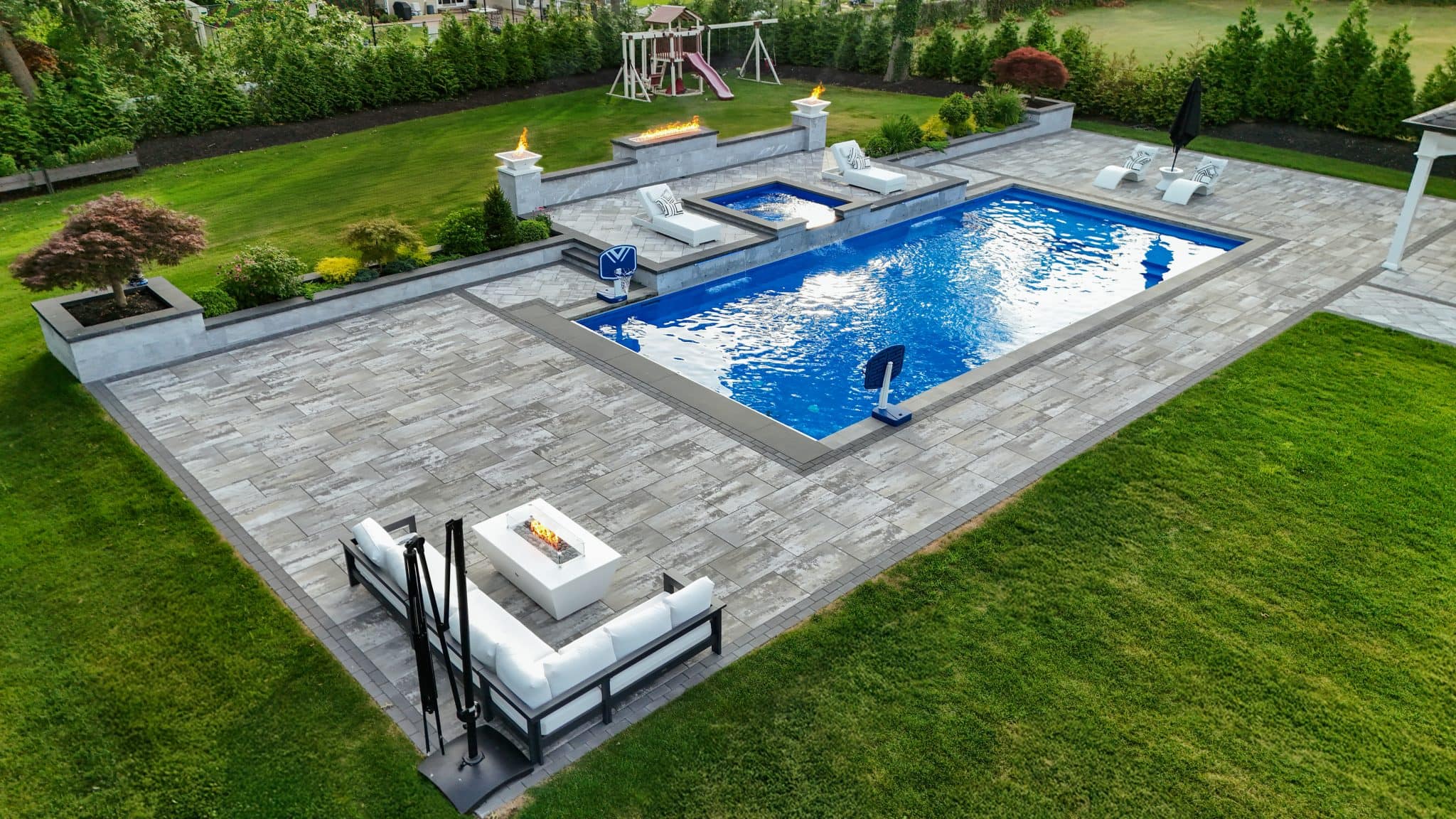 Modern backyard with rectangular swimming pool, lounge seating, fire features, and manicured lawn; playset visible in the background near trees.