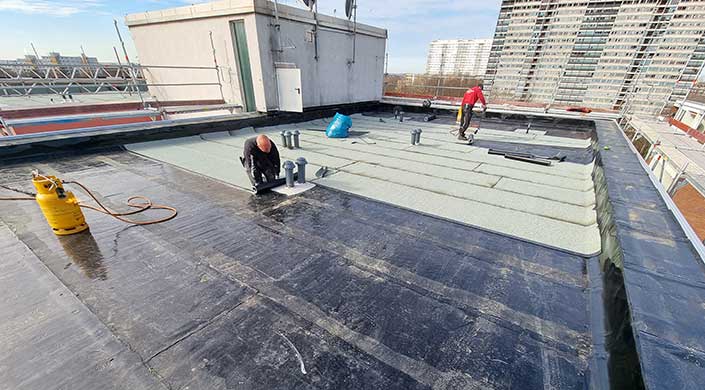 Flachdach mit mehreren Lüftungsrohren, einer blauen Plastiktüte und einem weißen Technikgebäude im Hintergrund bei Tageslicht.