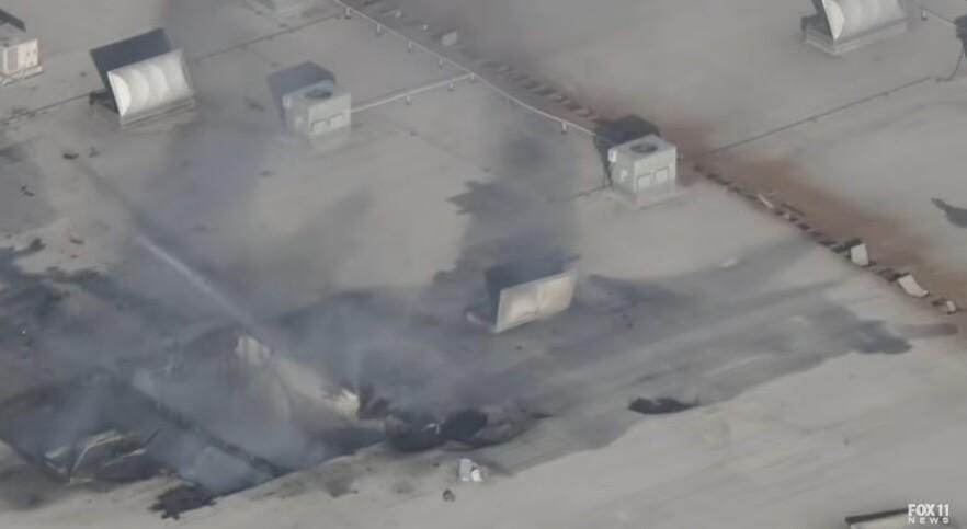 Aerial view of a warehouse roof after a plane crash in Southern California, with smoke rising from a large hole. Firefighters are likely responding to a fire. The plane collided with the warehouse, creating a large hole in the roof. Evidence from crash investigation.