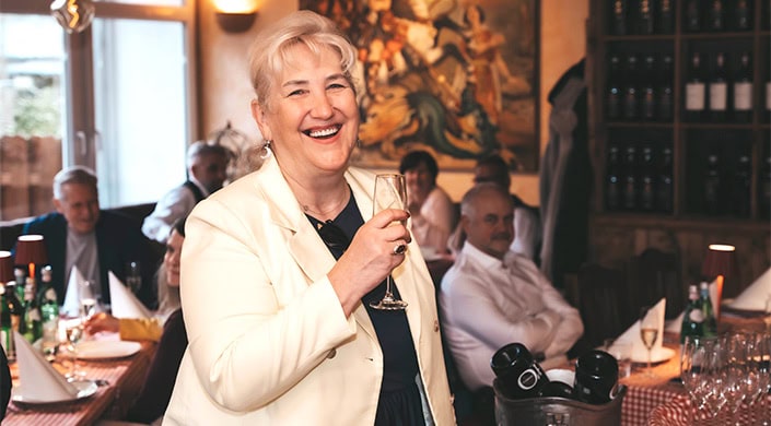 Ältere Frau mit weißem Haar lächelt und hält ein Glas Sekt in der Hand, im Hintergrund sitzen weitere Personen in einem festlichen Raum.
