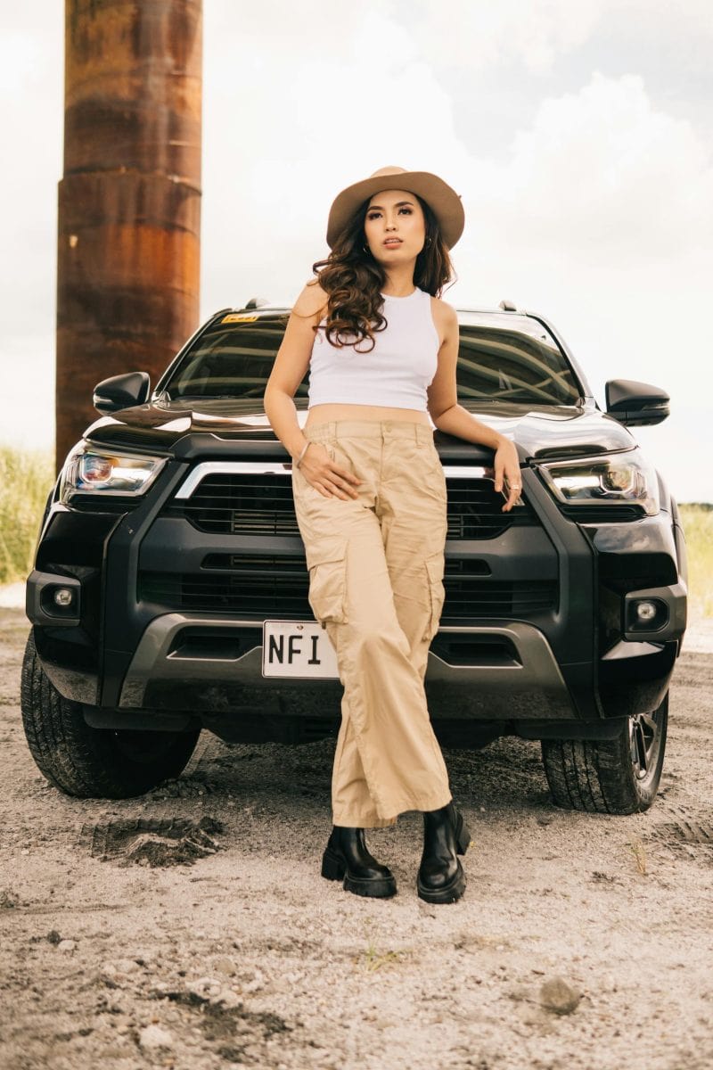 Fashionable woman in a hat and cargo pants posing confidently against a black SUV on a sunny day.