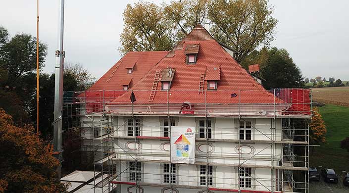 Ein Haus mit rotem Dach, Gerüst und Bauplakat, Leitern an der Fassade, Bäume im Hintergrund.