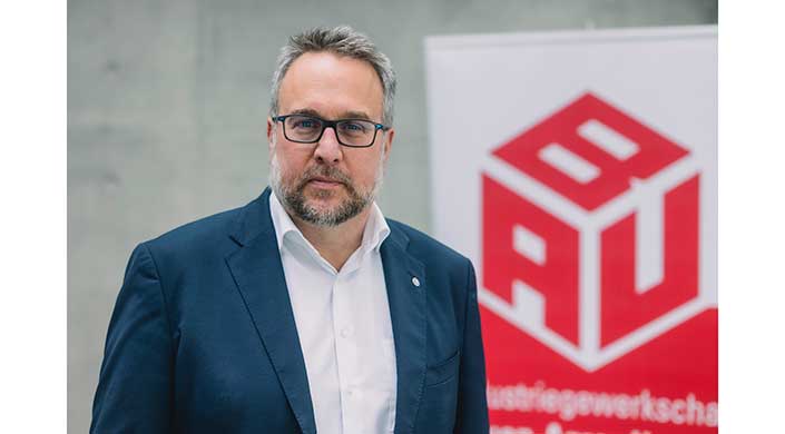 Ein Mann mit Brille, Bart und grauen Haaren trägt einen dunklen Anzug und weißes Hemd, steht vor einem unscharfen Hintergrund drinnen.
