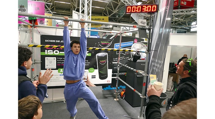Messestand mit Werbeplakat für einen schwarzen Zylinderbehälter, Gerüste aus Metall, Teil einer Person im Vordergrund, Hallendecke sichtbar.