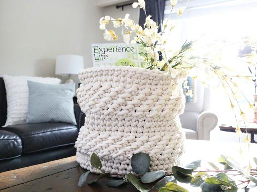 A thick, textured white basket on a table contains a magazine and white flowers; in the softly lit background, there is a couch with pillows and a lamp. Free crochet pattern