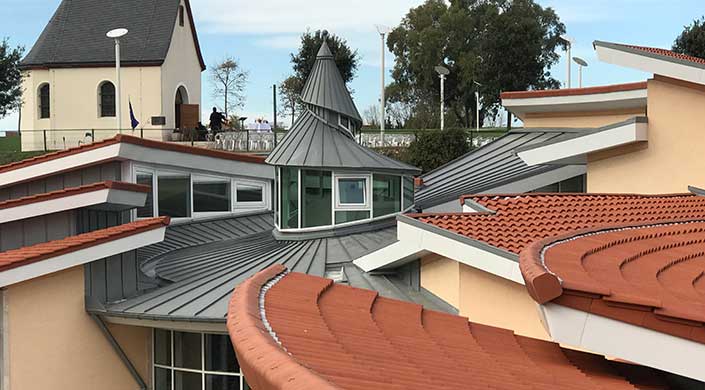 Modernes Gebäude mit rundem Turm, Metallverkleidung und vielen Fenstern. Im Vordergrund rote Ziegeldächer, im Hintergrund Bäume und Himmel.