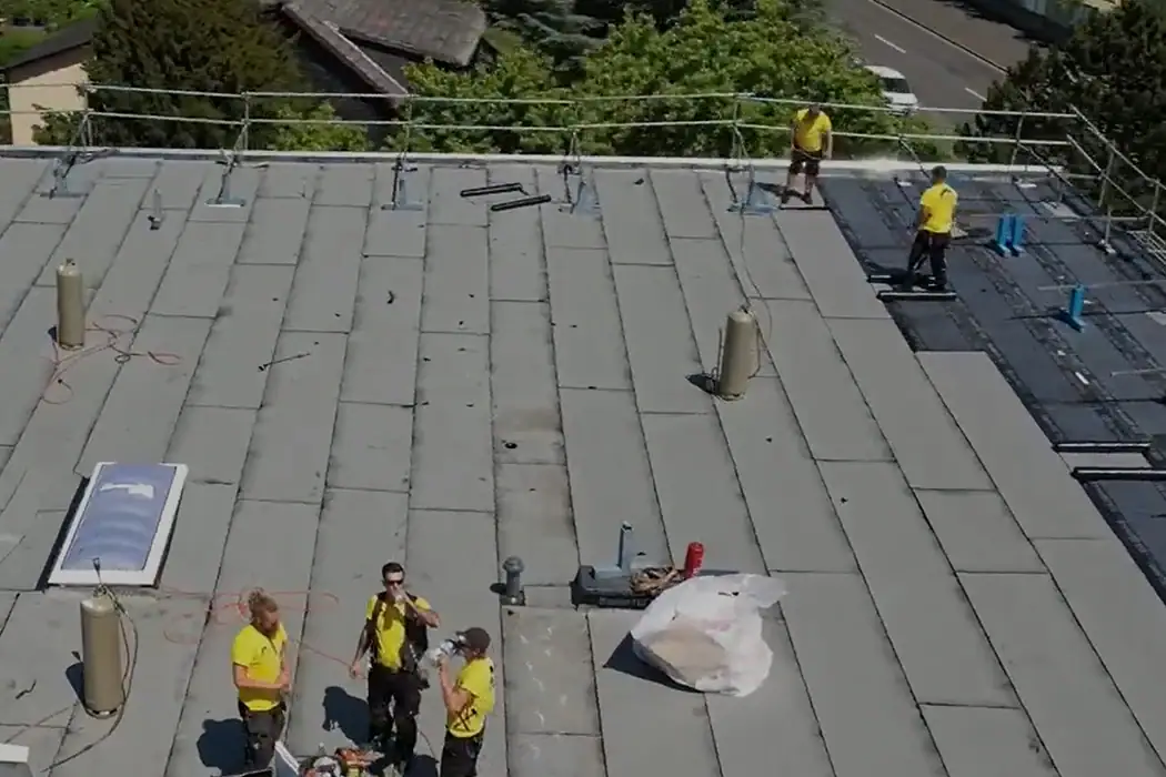 Mehrere Dachdecker in gelben Hemden arbeiten und pausieren auf einem Flachdach mit Sicherheitsgeländer und Baumaterialien, im Hintergrund Bäume und eine Straße.