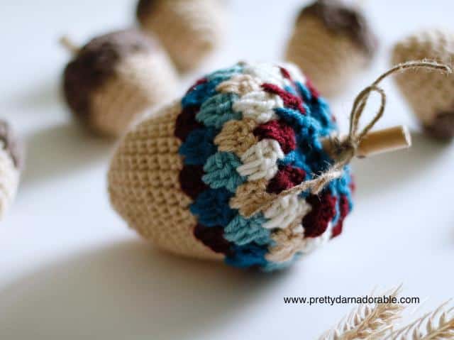 A beige-based crocheted acorn with a blue, red, and beige cap tied with twine rests among similar acorns on a white surface. Free crochet pattern