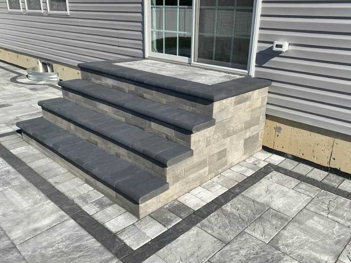 Gray stone steps with dark stone edges leading to a door, surrounded by a matching stone patio and light gray siding.