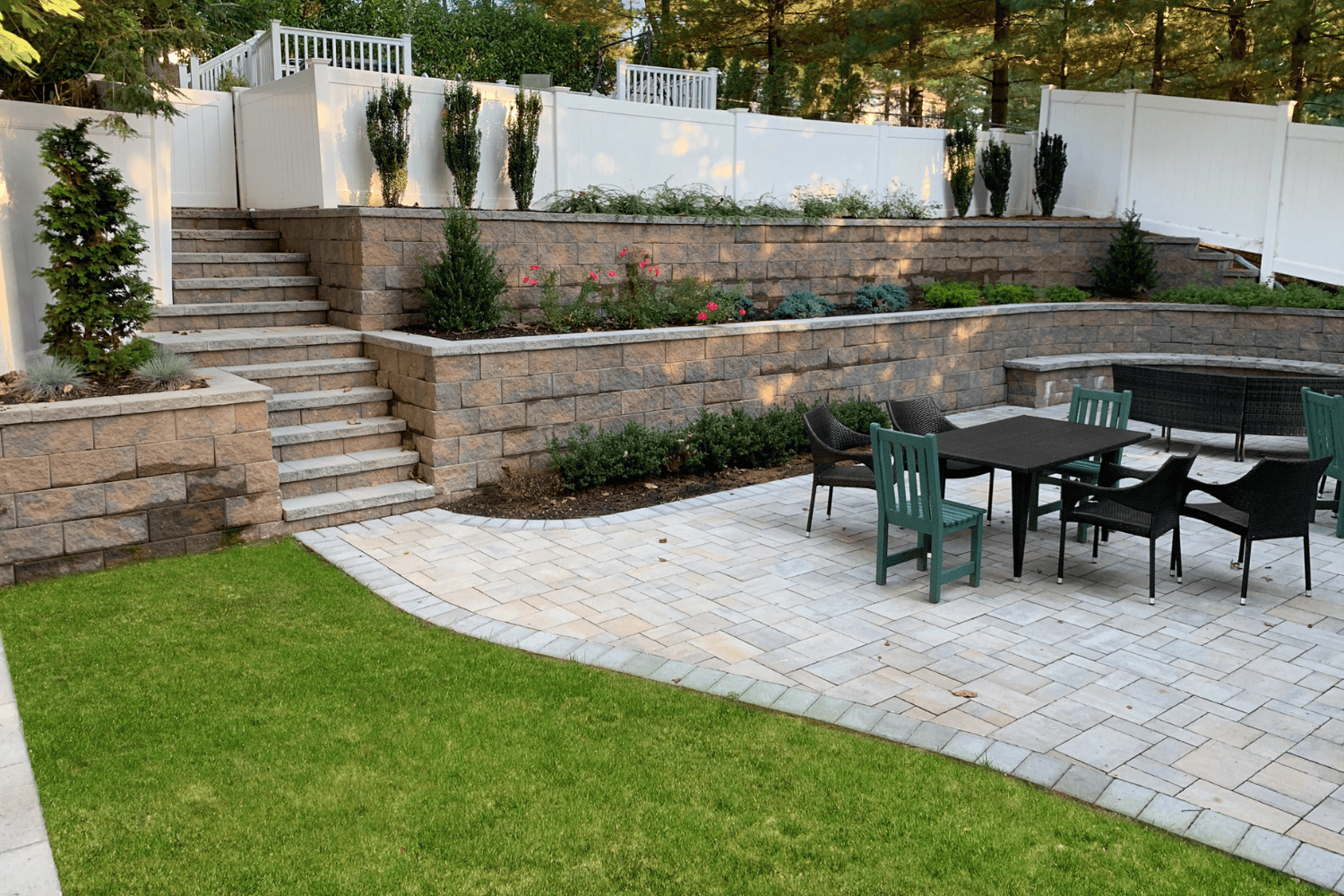 A backyard patio with a dining table and chairs on stone pavers, bordered by elegant retaining walls, steps, green lawn, and landscaped plants.