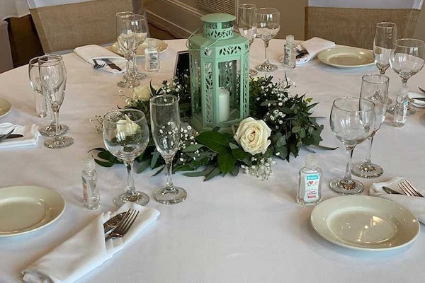 A round table set for a formal event with glassware, plates, silverware, napkins, hand sanitizers, and a floral centerpiece with a lantern and candle.