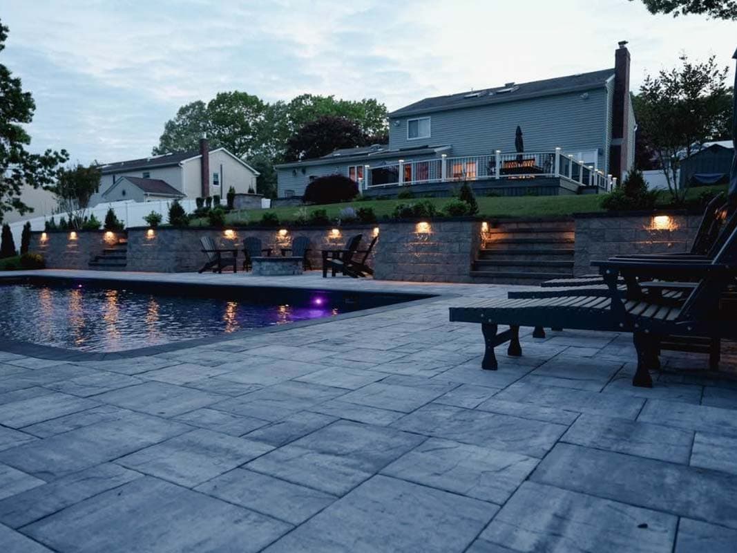Backyard with a swimming pool bordered by a retaining wall, lounge chairs, and a two-story house in the background. The patio area has stone tiles, and there are steps leading up to the elevated deck with outdoor seating in Elwood.