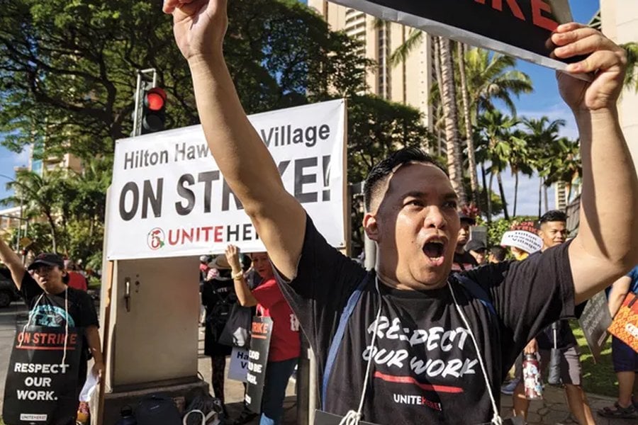 A man passionately protesting during a labor strike in front of a banner reading "Hilton Hawaiian Village ON STRIKE! UNITE HERE." The man is wearing a black T-shirt with the text "RESPECT OUR WORK" and raising his fist in the air, expressing determination and solidarity. Other protesters in the background hold similar signs, with tropical trees and buildings visible under a clear sky.