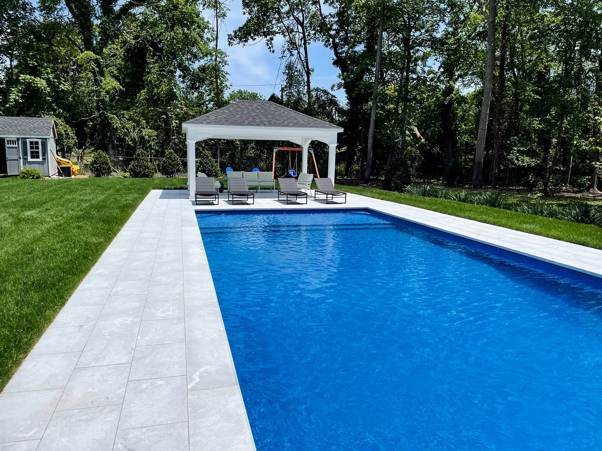 Rectangular outdoor swimming pool with a white gazebo and lounge chairs on a stone patio, surrounded by grass and trees on a sunny day.