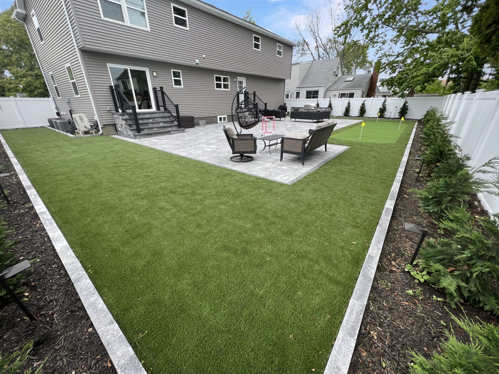 A backyard with artificial grass, a paved patio area with outdoor furniture, a fire pit, and a swing. There are shrubs along the fence and a small putting green.