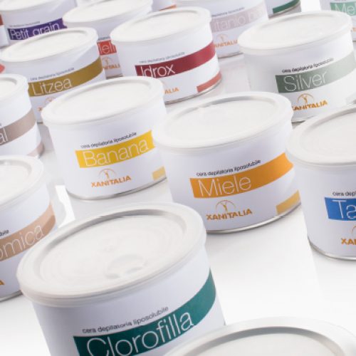 Diverse potten ontharingswax van Xanitalia, elk gelabeld met verschillende geuren en ingrediënten zoals honing, banaan en talkpoeder.