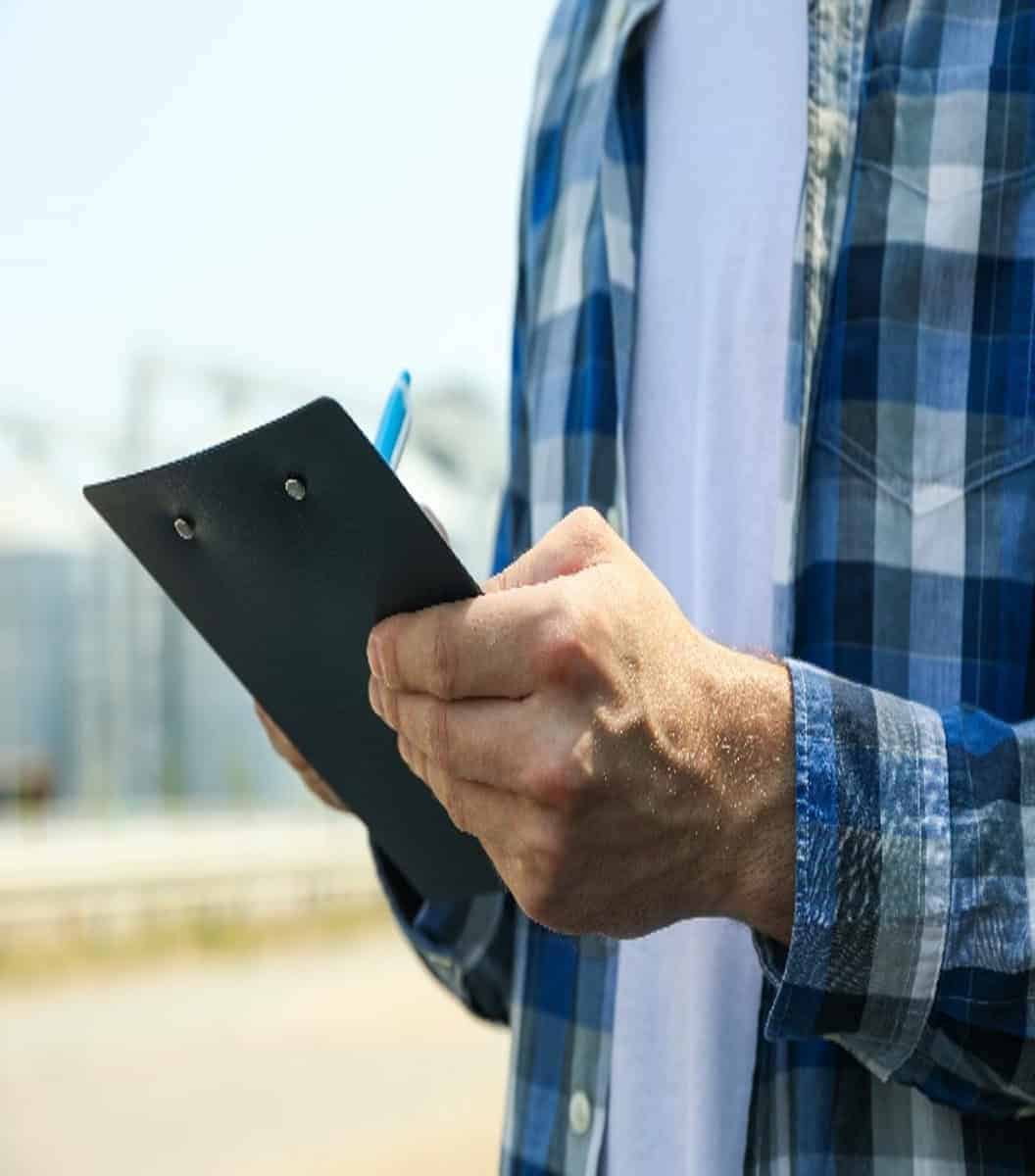 A person wearing a plaid shirt holds a clipboard and pen, standing outdoors with blurred industrial buildings in the background.