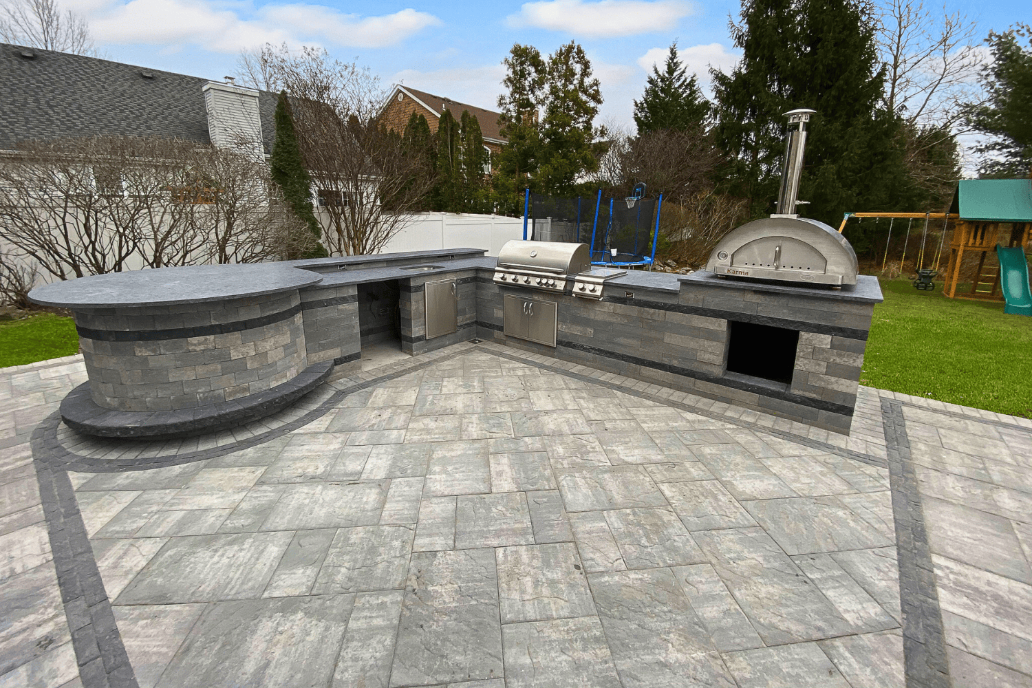 Outdoor Kitchens elevate your backyard with stone countertops, a built-in grill, pizza oven, and storage compartments on a paved patio, all surrounded by grass and trees.